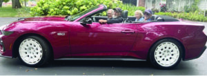 Four people ride together in a burgundy convertible classic car with white-wall tires, driving through a tree-lined residential street
