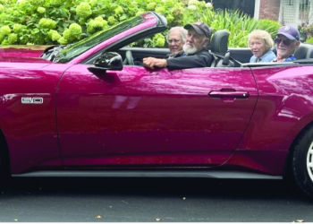 Four people ride together in a burgundy convertible classic car with white-wall tires, driving through a tree-lined residential street