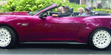 Four people ride together in a burgundy convertible classic car with white-wall tires, driving through a tree-lined residential street