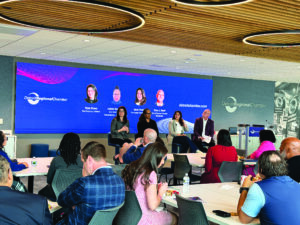 A panel discussion at the Detroit Regional Chamber features four speakers seated in front of a large blue screen displaying their names and titles. The audience sits at tables in the foreground, with the Detroit Regional Chamber logo and event branding visible on the backdrop