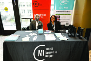 Two professionals sit at a table branded with "MI small business helper" logos at an event booth. Behind them are banners for the New Economy Initiative and the small business helper program, with promotional materials and merchandise displayed on the table in front of them
