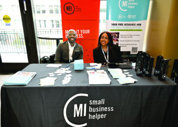 Two professionals sit at a table branded with "MI small business helper" logos at an event booth. Behind them are banners for the New Economy Initiative and the small business helper program, with promotional materials and merchandise displayed on the table in front of them