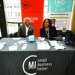 Two professionals sit at a table branded with "MI small business helper" logos at an event booth. Behind them are banners for the New Economy Initiative and the small business helper program, with promotional materials and merchandise displayed on the table in front of them