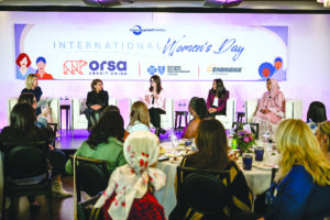 A panel discussion during an International Women's Day event hosted by the Detroit Regional Chamber, featuring five women panelists on stage with logos from sponsor organizations including Orsa Credit Union, Blue Cross Blue Shield, and Enbridge. The audience is seated at tables in the foreground.