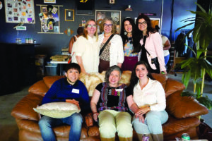 A group of eight people poses together for a photo—four standing in back and four seated on a brown leather couch in front—at an indoor event space with dark walls decorated with framed photos, artwork, and potted plants visible in the background