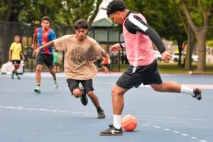 A young player in a tan Arizona graphic shirt dribbles an orange soccer ball while being defended by a player in a pink and black vest on a blue and pink court, with other youth participants and trees visible in the background