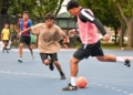 A young player in a tan Arizona graphic shirt dribbles an orange soccer ball while being defended by a player in a pink and black vest on a blue and pink court, with other youth participants and trees visible in the background
