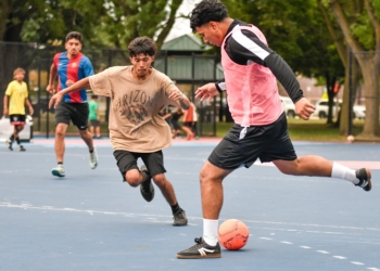 A young player in a tan Arizona graphic shirt dribbles an orange soccer ball while being defended by a player in a pink and black vest on a blue and pink court, with other youth participants and trees visible in the background