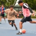 A young player in a tan Arizona graphic shirt dribbles an orange soccer ball while being defended by a player in a pink and black vest on a blue and pink court, with other youth participants and trees visible in the background