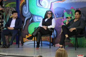 Three panelists sit on stage during a discussion—a man in a navy blazer on the left, a woman in the center wearing glasses and a pinstriped vest, and a man on the right in dark clothing—with a colorful backdrop featuring vibrant green, turquoise, and purple artwork depicting urban and floral imagery