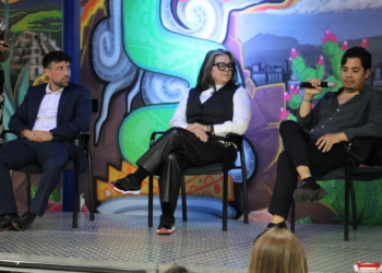 Three panelists sit on stage during a discussion—a man in a navy blazer on the left, a woman in the center wearing glasses and a pinstriped vest, and a man on the right in dark clothing—with a colorful backdrop featuring vibrant green, turquoise, and purple artwork depicting urban and floral imagery