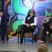 Three panelists sit on stage during a discussion—a man in a navy blazer on the left, a woman in the center wearing glasses and a pinstriped vest, and a man on the right in dark clothing—with a colorful backdrop featuring vibrant green, turquoise, and purple artwork depicting urban and floral imagery