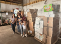 A family group—an adult woman, adult man, two children, and a small child—poses together in the warehouse standing in front of tall stacks of cardboard boxes containing food relief supplies