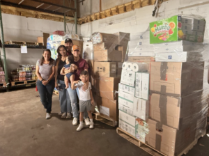 A family group—an adult woman, adult man, two children, and a small child—poses together in the warehouse standing in front of tall stacks of cardboard boxes containing food relief supplies