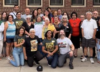 A large diverse group of El Colectivo Summer Gathering celebrants—alumni, staff, and friends representing multiple generations—pose together in front of a brick building for a group photo