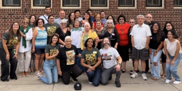 A large diverse group of El Colectivo Summer Gathering celebrants—alumni, staff, and friends representing multiple generations—pose together in front of a brick building for a group photo