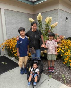 A family group stands together outdoors in front of a brick building with flowering plants—an adult woman with three children and an infant in a stroller, all smiling at the camera with yellow and reddish flowers blooming in the landscaped area behind them