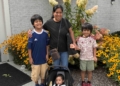 A family group stands together outdoors in front of a brick building with flowering plants—an adult woman with three children and an infant in a stroller, all smiling at the camera with yellow and reddish flowers blooming in the landscaped area behind them
