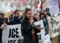 A woman speaks passionately into a microphone at an immigration rights protest, addressing a gathered crowd holding signs with messages about ICE, with bare winter trees and brick buildings visible in the background