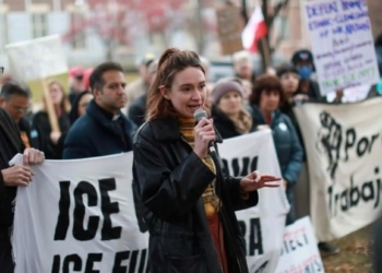 A woman speaks passionately into a microphone at an immigration rights protest, addressing a gathered crowd holding signs with messages about ICE, with bare winter trees and brick buildings visible in the background