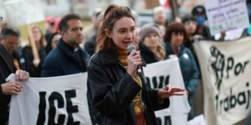 A woman speaks passionately into a microphone at an immigration rights protest, addressing a gathered crowd holding signs with messages about ICE, with bare winter trees and brick buildings visible in the background