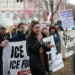 A woman speaks passionately into a microphone at an immigration rights protest, addressing a gathered crowd holding signs with messages about ICE, with bare winter trees and brick buildings visible in the background