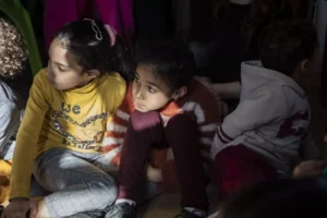 Two young children sit together at what appears to be a community event or gathering, one wearing a yellow shirt and the other in maroon-colored clothing, with adults visible partially in the frame around them