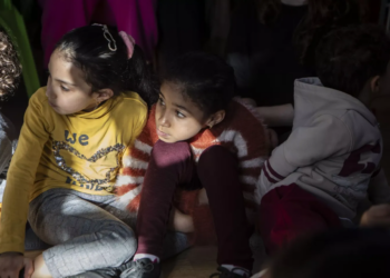 Two young children sit together at what appears to be a community event or gathering, one wearing a yellow shirt and the other in maroon-colored clothing, with adults visible partially in the frame around them
