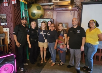 A group of seven people stand together in front of a presentation screen inside the venue, with the yellow soccer ball visible and neon speakers and event decor in the background