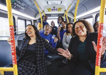 A diverse group of approximately twelve people celebrate inside a Detroit public transit bus (numbered 2527). Two women are prominently featured in the foreground—one wearing a black and white plaid blazer on the left, the other in a black blazer with glasses on the right—both smiling and gesturing. Additional passengers stand and sit throughout the bus behind them, several wearing blue clothing. Yellow handrails and support poles frame the scene, with red banners visible that read "Believe Your Story." Windows show the street outside, and the interior has blue-gray seating.
