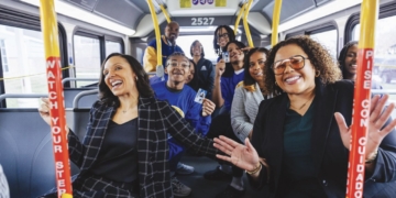 A diverse group of approximately twelve people celebrate inside a Detroit public transit bus (numbered 2527). Two women are prominently featured in the foreground—one wearing a black and white plaid blazer on the left, the other in a black blazer with glasses on the right—both smiling and gesturing. Additional passengers stand and sit throughout the bus behind them, several wearing blue clothing. Yellow handrails and support poles frame the scene, with red banners visible that read "Believe Your Story." Windows show the street outside, and the interior has blue-gray seating.