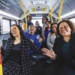 A diverse group of approximately twelve people celebrate inside a Detroit public transit bus (numbered 2527). Two women are prominently featured in the foreground—one wearing a black and white plaid blazer on the left, the other in a black blazer with glasses on the right—both smiling and gesturing. Additional passengers stand and sit throughout the bus behind them, several wearing blue clothing. Yellow handrails and support poles frame the scene, with red banners visible that read "Believe Your Story." Windows show the street outside, and the interior has blue-gray seating.
