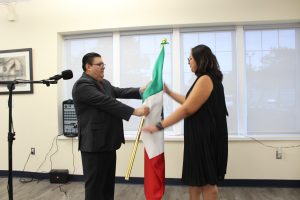 Man in dark suit with glasses and red tie speaking at microphone next to woman in black sleeveless dress. Both holding and presenting large Mexican flag together. Large windows with daylight in background. Framed artwork on cream-colored walls. Official government office setting.