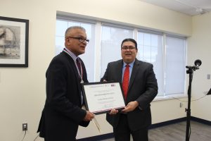 Two men in dark suits, one holding a framed award certificate labeled "RECONOCIMIENTO." Man on left wearing a maroon tie and medal on ribbon. Formal office setting with windows.