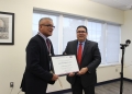 Two men in dark suits, one holding a framed award certificate labeled "RECONOCIMIENTO." Man on left wearing a maroon tie and medal on ribbon. Formal office setting with windows.
