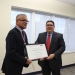 Two men in dark suits, one holding a framed award certificate labeled "RECONOCIMIENTO." Man on left wearing a maroon tie and medal on ribbon. Formal office setting with windows.