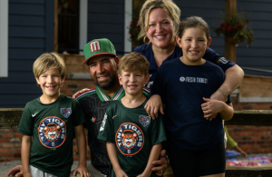 Family portrait of four people outdoors smiling together. Man in center wears a green and red Mexican flag-inspired baseball cap and casual shirt, with his arm around a woman in a dark navy "FIESTA TIGRES" shirt. Two young boys in front wear matching green Detroit Tigers logo t-shirts. All four are smiling warmly at the camera. White siding of house visible in background. Casual outdoor family moment captured in daylight.