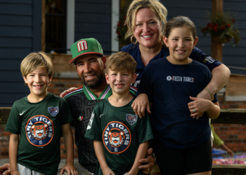 Family portrait of four people outdoors smiling together. Man in center wears a green and red Mexican flag-inspired baseball cap and casual shirt, with his arm around a woman in a dark navy "FIESTA TIGRES" shirt. Two young boys in front wear matching green Detroit Tigers logo t-shirts. All four are smiling warmly at the camera. White siding of house visible in background. Casual outdoor family moment captured in daylight.