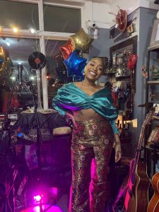 Woman smiling at camera indoors in a vibrant creative studio space. She wears a turquoise silk shawl draped over her shoulders and burgundy-and-gold patterned pants with geometric designs. Colorful foil star balloons in red, gold, and blue hang from the ceiling. Purple and magenta LED lighting illuminates the space. Guitars and other musical instruments visible on the right side. Large windows with white frames in background. Decorative items, shelving, and holiday ornaments scattered throughout the creative workspace. Festive and energetic atmosphere.