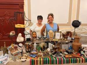 Two women posing behind a table displaying curated Mexican and home décor items in an elegant formal space. Woman on left wears cream-colored top with black glasses and dark bangs. Woman on right wears light blue sleeveless dress with gold earrings and upswept hair. Table displays brass candlesticks, beaded necklaces, woven baskets, decorative plates with Mexican patterns, folk art masks, bowls, and cultural artifacts. Ornate red-burgundy door with decorative molding and cream walls visible behind them. Black tiered stand with yellow bowl on left side
