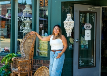 Woman with long brown hair in a top bun indoors, wearing a black v-neck top and blue jeans with brown braided belt. She's smiling brightly at the camera with both hands raised playfully near her head. Gold chain necklaces and rings visible. Warm golden lighting illuminates the space, with shelving and home décor items blurred in the background