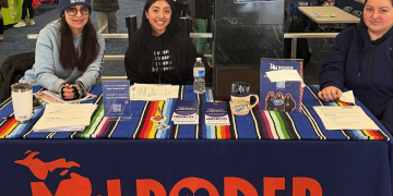 Three women seated at a table branded "MI PODER" during an event or conference, with colorful striped table runners and informational materials, with event attendees visible in the background.