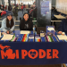 Three women seated at a table branded "MI PODER" during an event or conference, with colorful striped table runners and informational materials, with event attendees visible in the background.