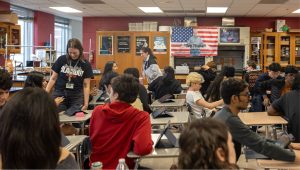Classroom scene with a teacher in a black shirt standing among students seated at desks with tablets. Burgundy walls display a US flag and educational posters in the background.