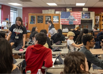 Classroom scene with a teacher in a black shirt standing among students seated at desks with tablets. Burgundy walls display a US flag and educational posters in the background.