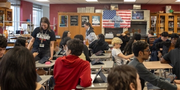 Classroom scene with a teacher in a black shirt standing among students seated at desks with tablets. Burgundy walls display a US flag and educational posters in the background.
