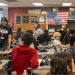 Classroom scene with a teacher in a black shirt standing among students seated at desks with tablets. Burgundy walls display a US flag and educational posters in the background.