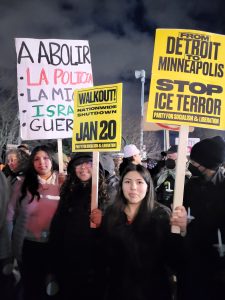 Nighttime protest scene with participants holding signs, including messages in Spanish about police abolition and signs reading "WALKOUT! NATIONWIDE SHUTDOWN JAN 20" and "FROM DETROIT TO MINNEAPOLIS STOP ICE TERROR." Participants bundled in winter clothing.