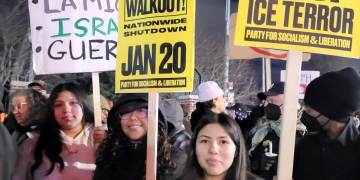 Nighttime protest scene with participants holding signs, including messages in Spanish about police abolition and signs reading "WALKOUT! NATIONWIDE SHUTDOWN JAN 20" and "FROM DETROIT TO MINNEAPOLIS STOP ICE TERROR." Participants bundled in winter clothing.