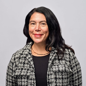 Professional headshot of a woman with black shoulder-length hair wearing a black and white patterned blazer over a black shirt against a neutral gray background.